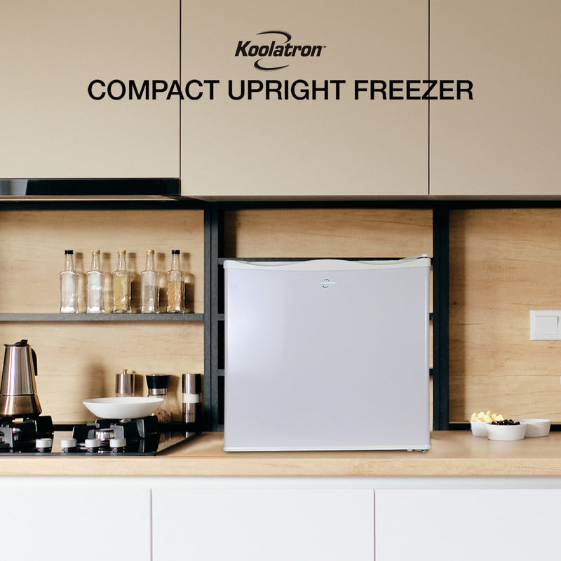 Lifestyle image of mini freezer on a light-colored wooden countertop with beige cupboards above and white cupboards below. To the left is a black cooktop with a coffee pot and bottles of flavoring syrups on a shelf above. Text above reads "Koolatron compact upright freezer"