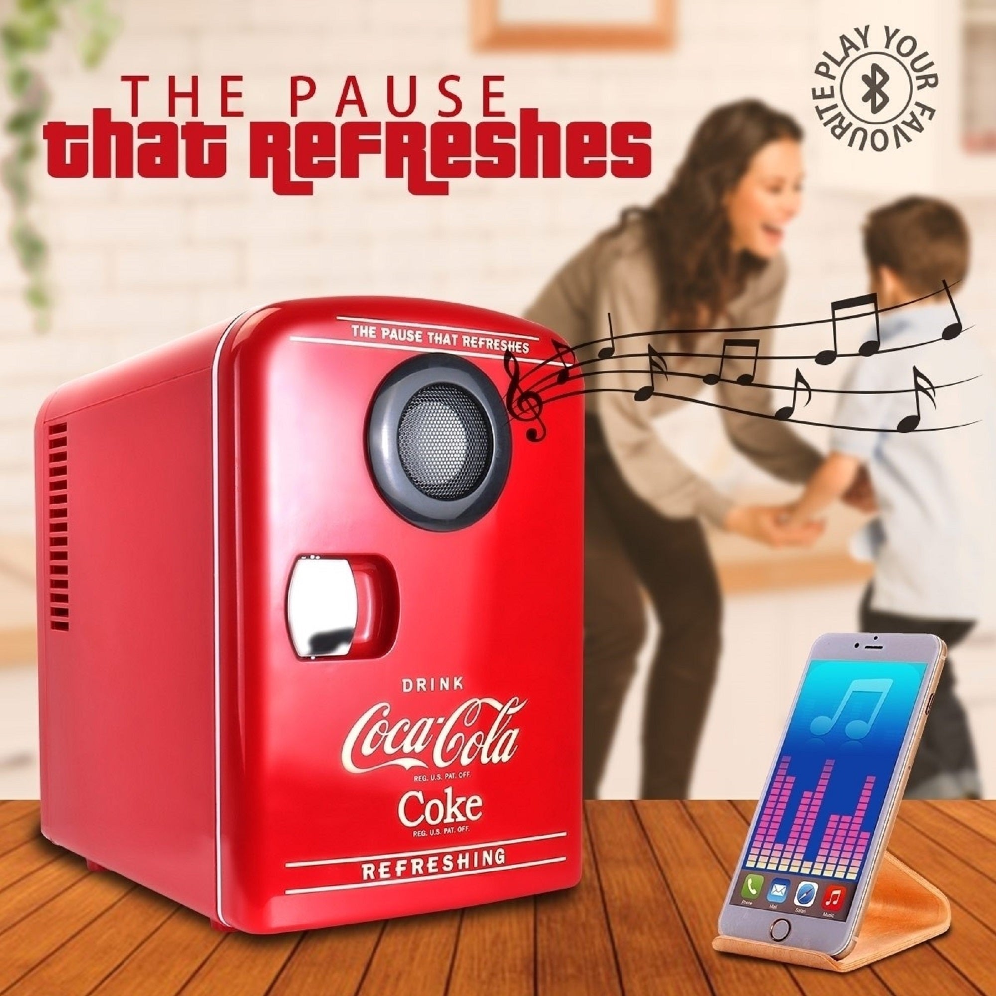 Lifestyle image of Coca-Cola 6 can cooler/wireless speaker beside a touchscreen music player on a wooden tabletop. An adult with long brown hair and a child with short brown hair are holding hands and dancing in the background. There are small black music notes coming out of the speaker and text above reads, "The pause that refreshes: Play your favorites"