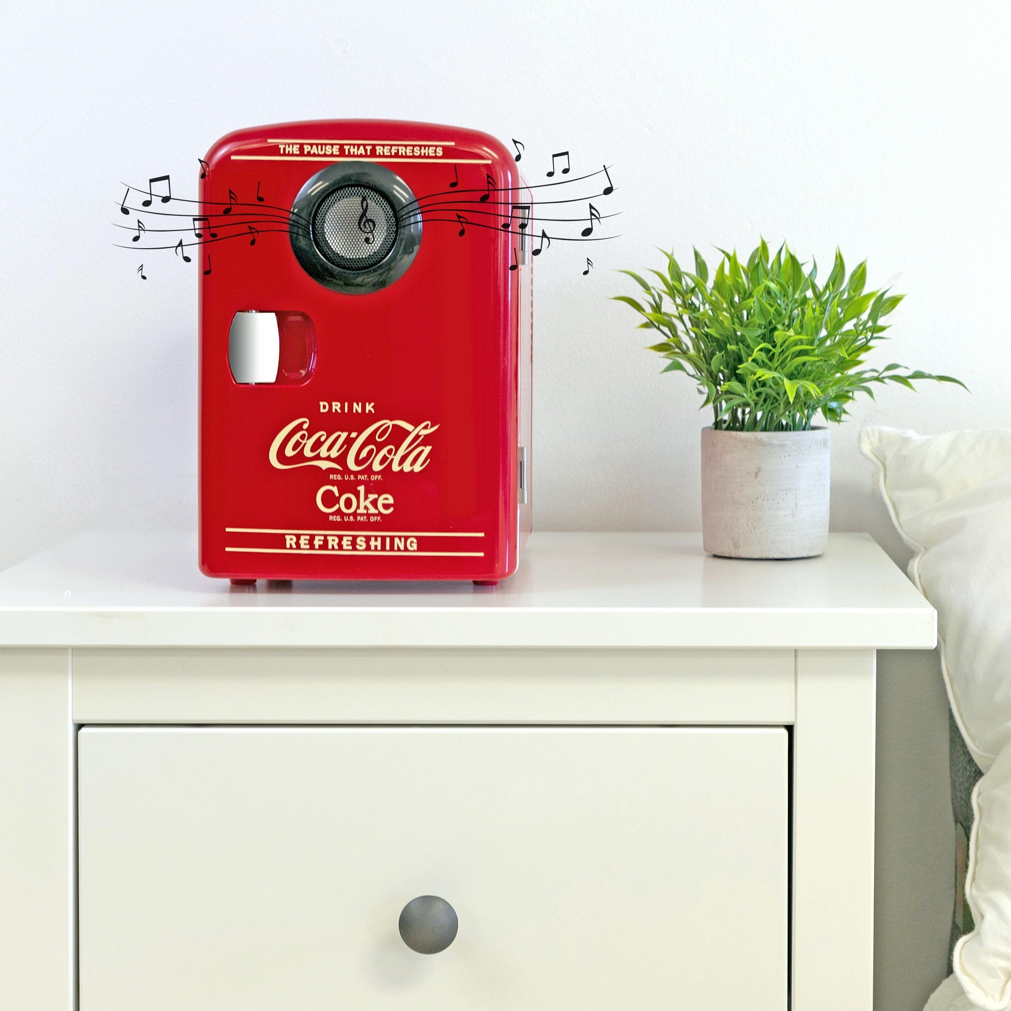 Lifestyle image of Coca-Cola 6 can mini fridge/speaker combo, closed, on a white bedside table with a plant in a white pot on its right, a white painted wall behind, and small black music notes overlaid on the speaker