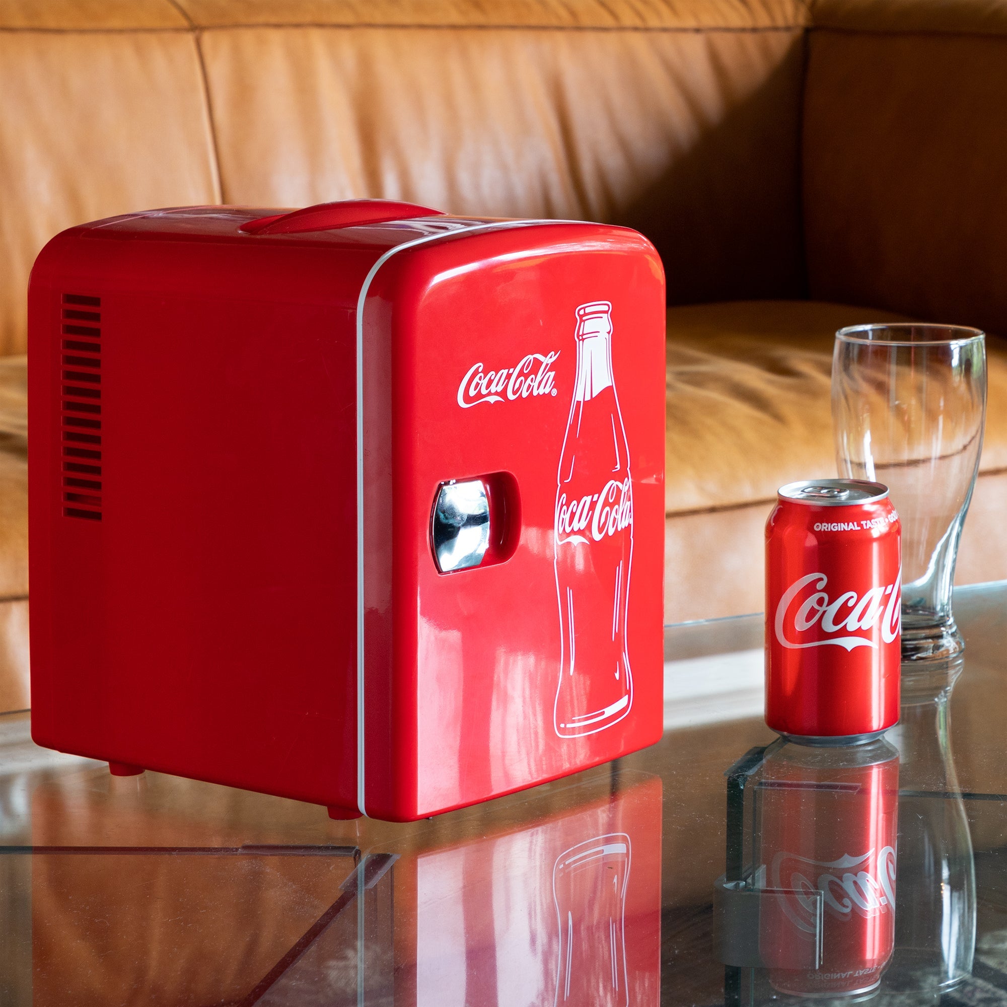 Lifestyle image of Coca-Cola Classic Bottle 6 can mini fridge, closed, on a glass coffee table with a tan leather sofa behind it and a can of Coke and empty glass to the right