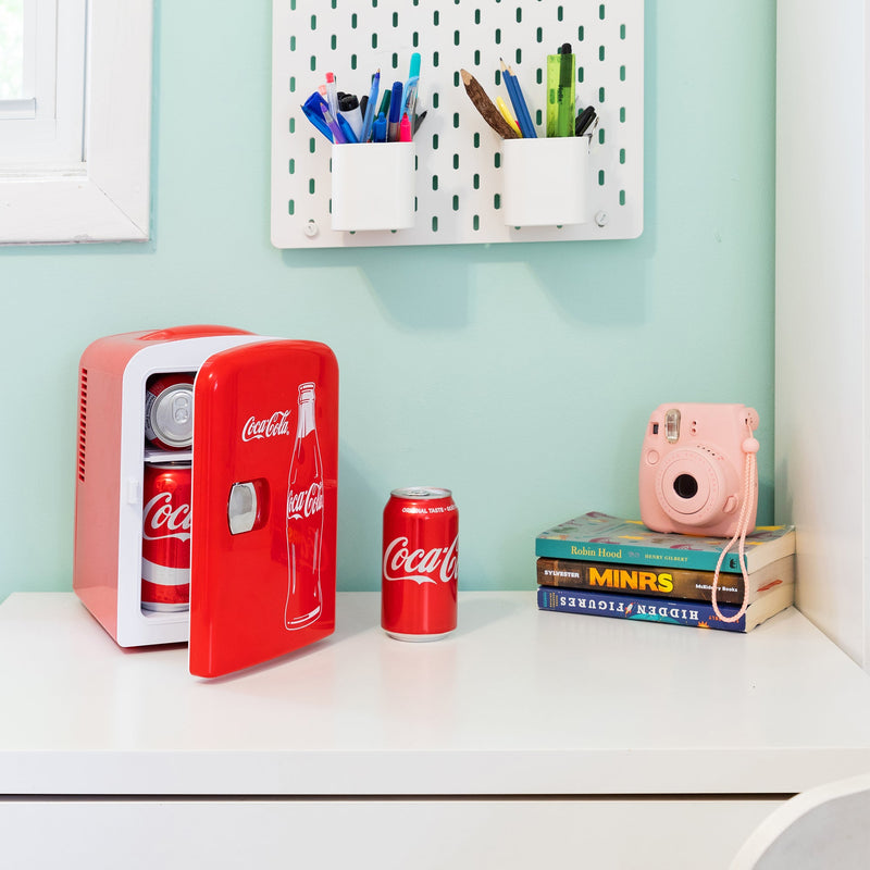 Lifestyle image of Coca-Cola Classic Bottle 6 can mini fridge, partly open with cans inside, on a white desktop with an aqua wall behind. There is a can of Coke, a stack of books, and a pink camera to the right of the fridge and two cups of pens and pencils attached to the wall above
