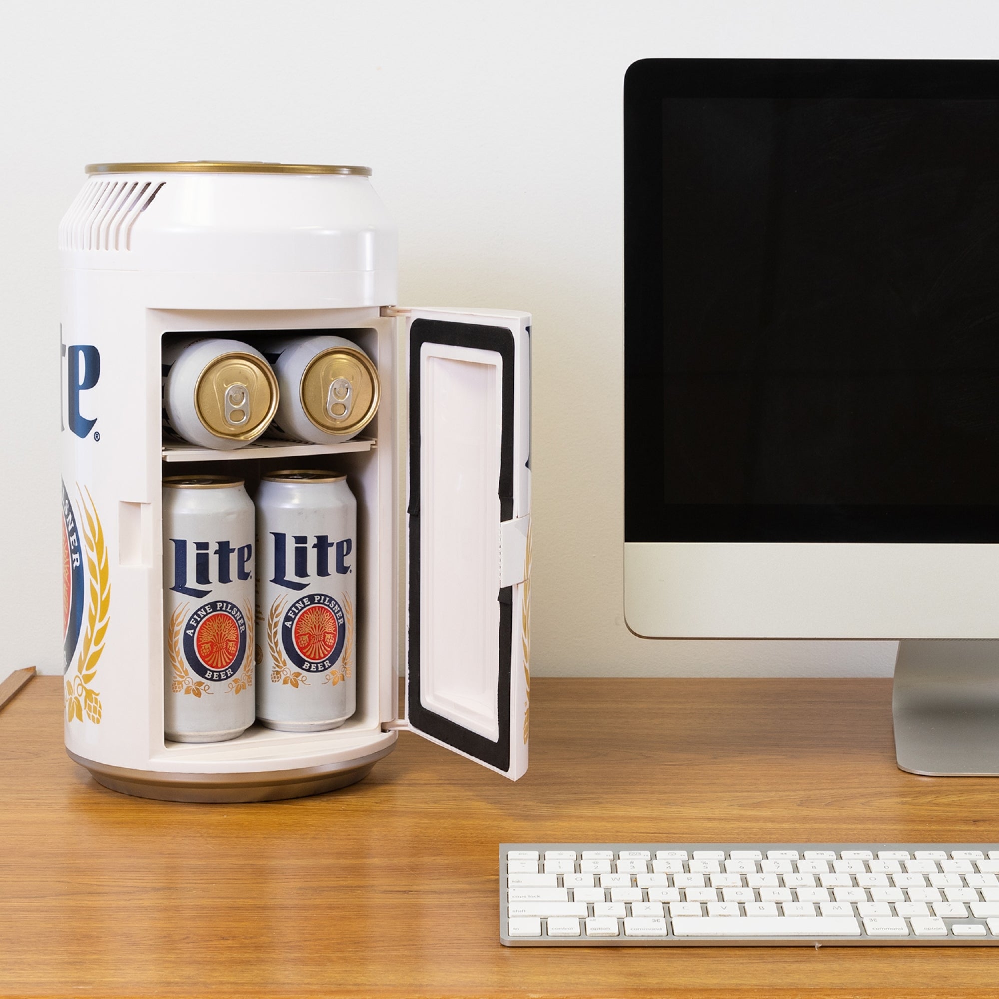 Lifestyle image of Miller Lite can-shaped mini fridge, open with 6 cans of Miller Lite beer inside, on a wooden desktop with a computer monitor and keyboard beside it
