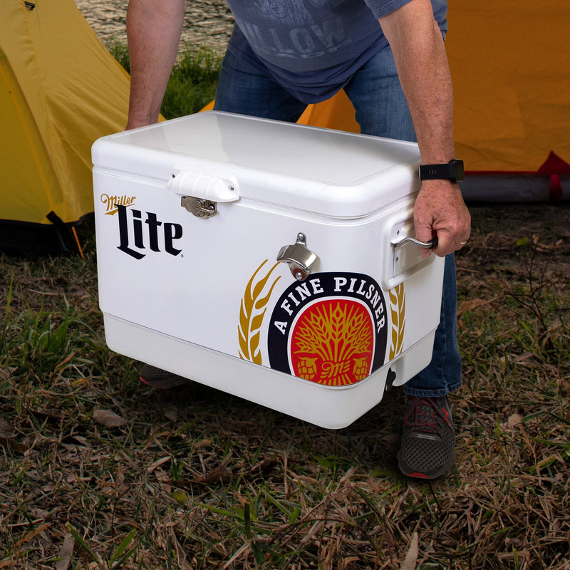 Lifestyle image of a person wearing jeans and a blue t-shirt lifting the Miller Lite 54 quart ice chest with two yellow dome tents visible in the background