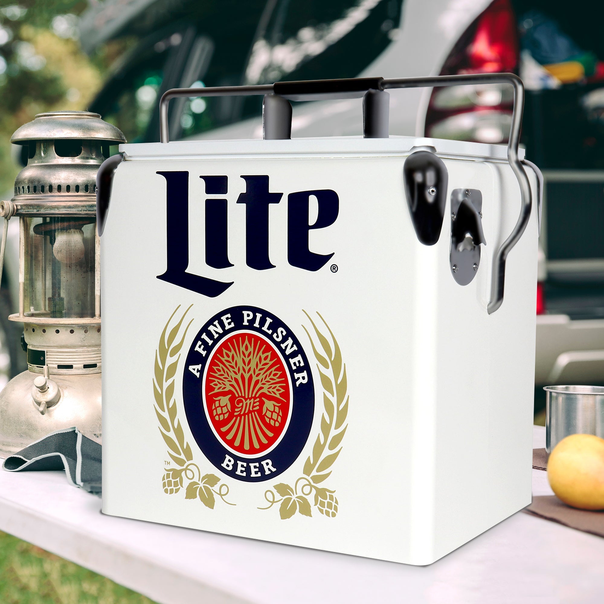 Lifestyle image of Miller Lite retro ice chest with bottle opener on a white folding table with a metal lantern beside it and a vehicle in the background