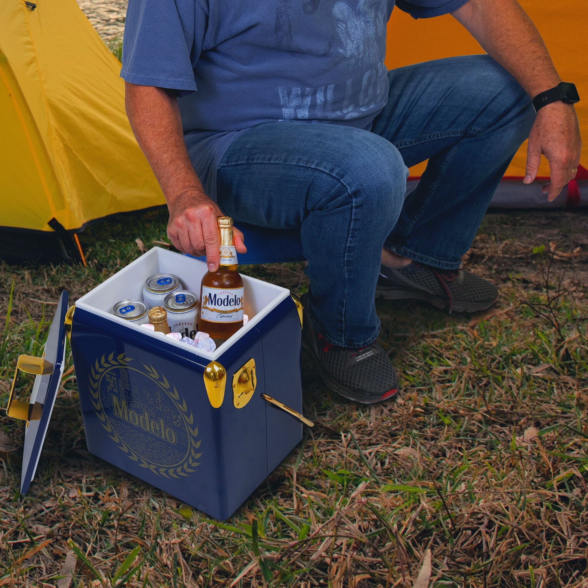 Lifestyle image of a person wearing jeans and a blue t-shirt sitting in front of two yellow dome tents and lifting a bottle of Modelo beer out of the open ice chest
