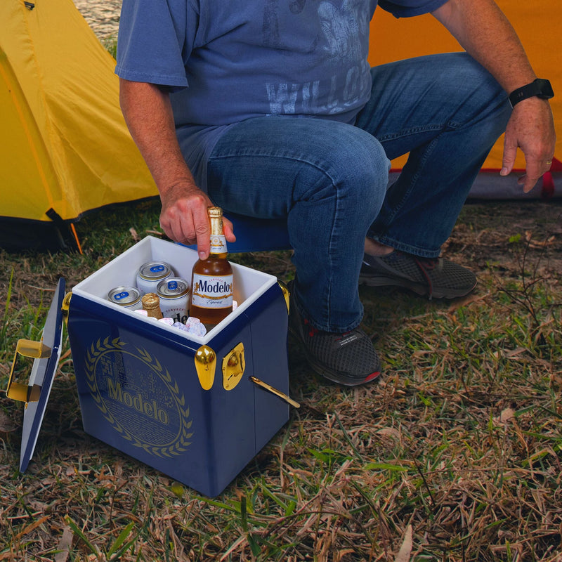 Lifestyle image of a person wearing jeans and a blue t-shirt sitting in front of two yellow dome tents and lifting a bottle of Modelo beer out of the open ice chest