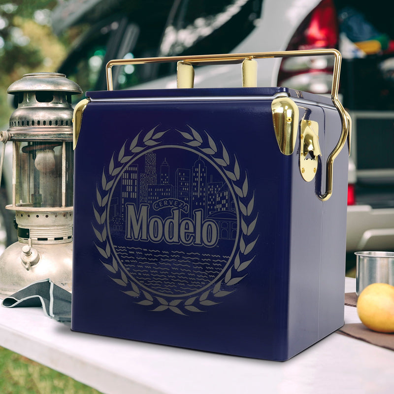 Lifestyle image of Modelo retro ice chest with bottle opener on a white folding table with a metal lantern beside it and a vehicle in the background