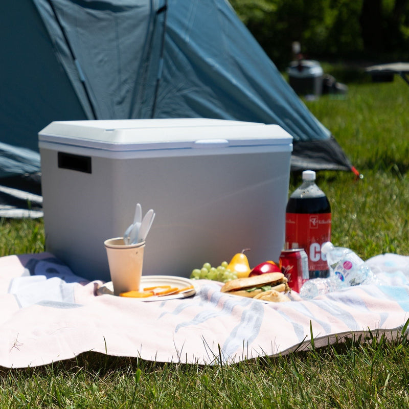 Lifestyle image of portable 12V cooler closed on a picnic blanket with food in front of it and a teal tent behind