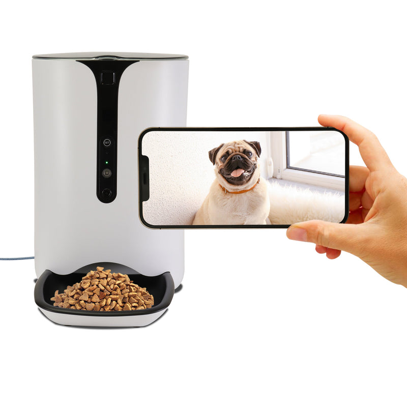 Product shot of Lentek smart pet feeder with dry pet food dispensed on a white background and a hand holding a smartphone with an image of a pug dog on the screen in front