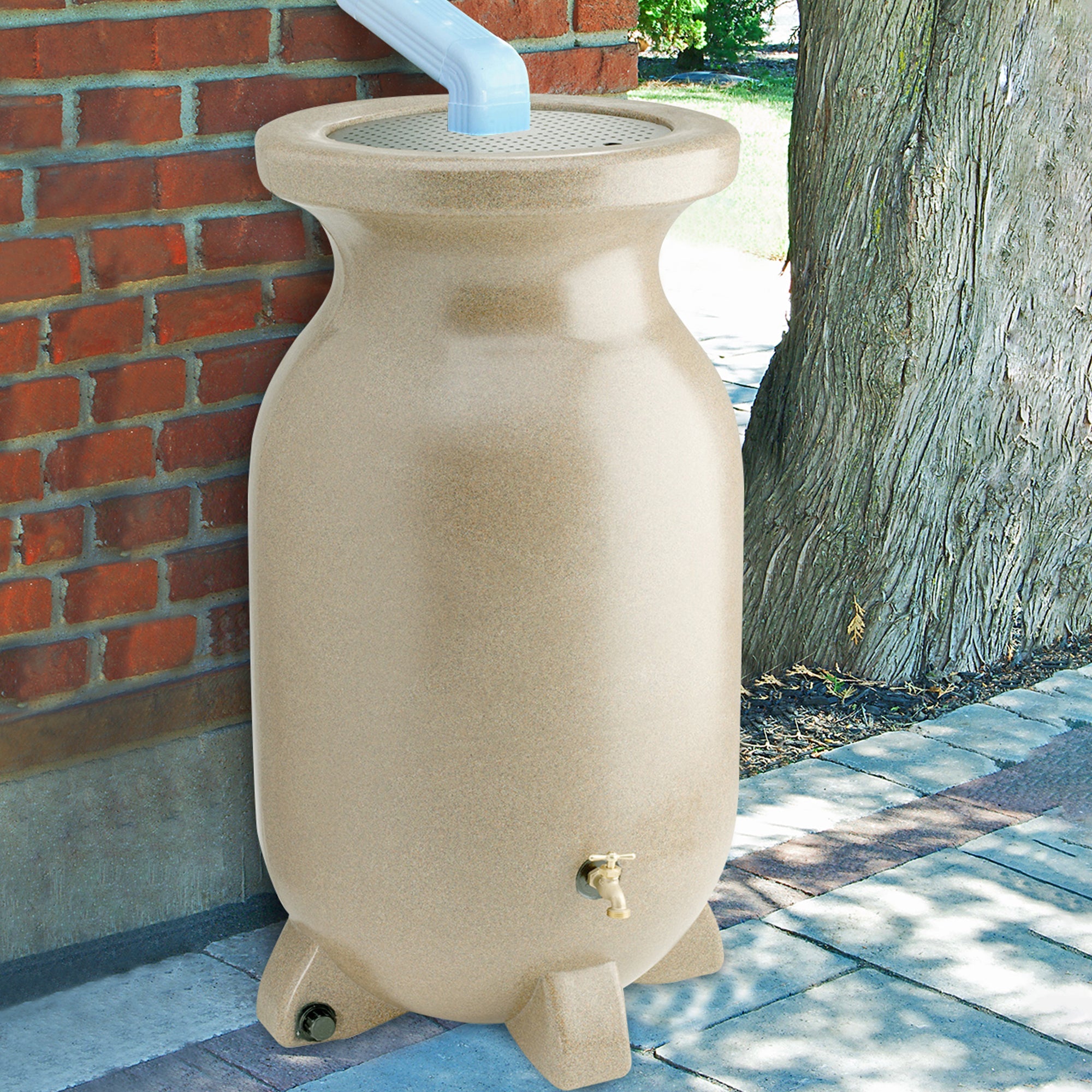 Lifestyle image of rain barrel set up on a brick pathway under a white downspout with a red brick wall and a tree in the background