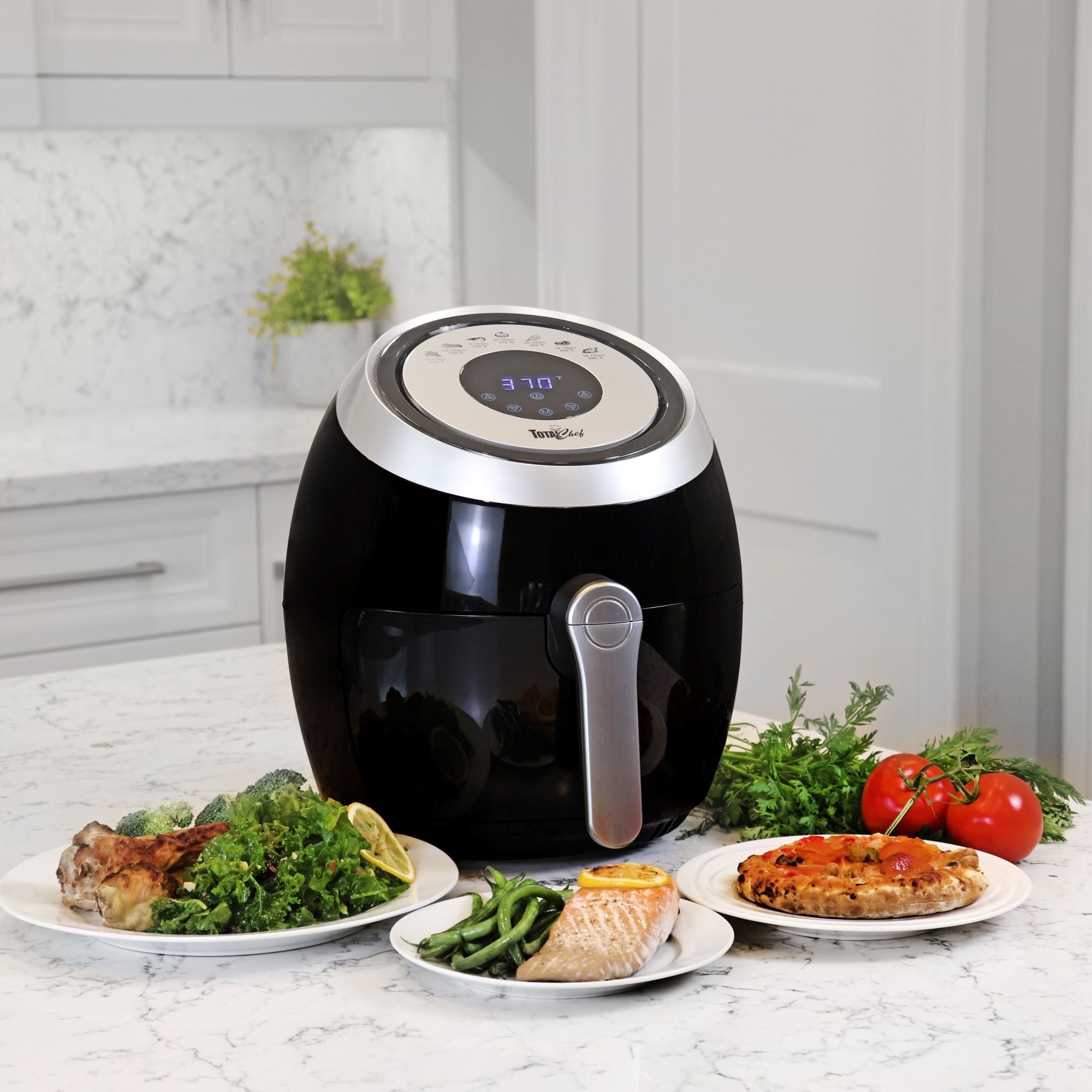 Lifestyle image of Total Chef air fryer on a white marble counter surrounded by plates of cooked food plus uncooked tomatoes and herbs