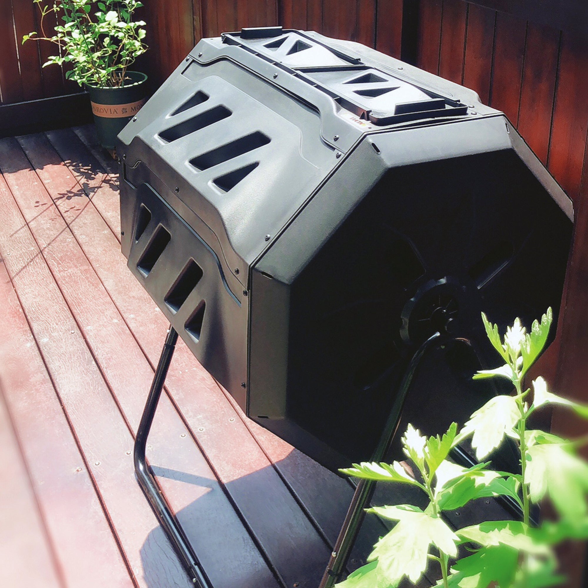 Lifestyle image of twin chamber tumbling composter set up on a red-stained wooden deck with potted plants on either side and a wooden fence behind it