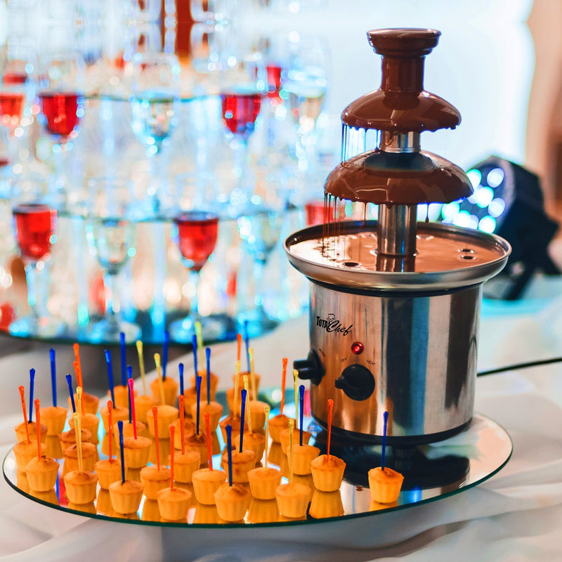 Lifestyle image of chocolate fountain filled with melted chocolate surrounded by mini cupcakes on skewers with a tower of wine glasses in the background