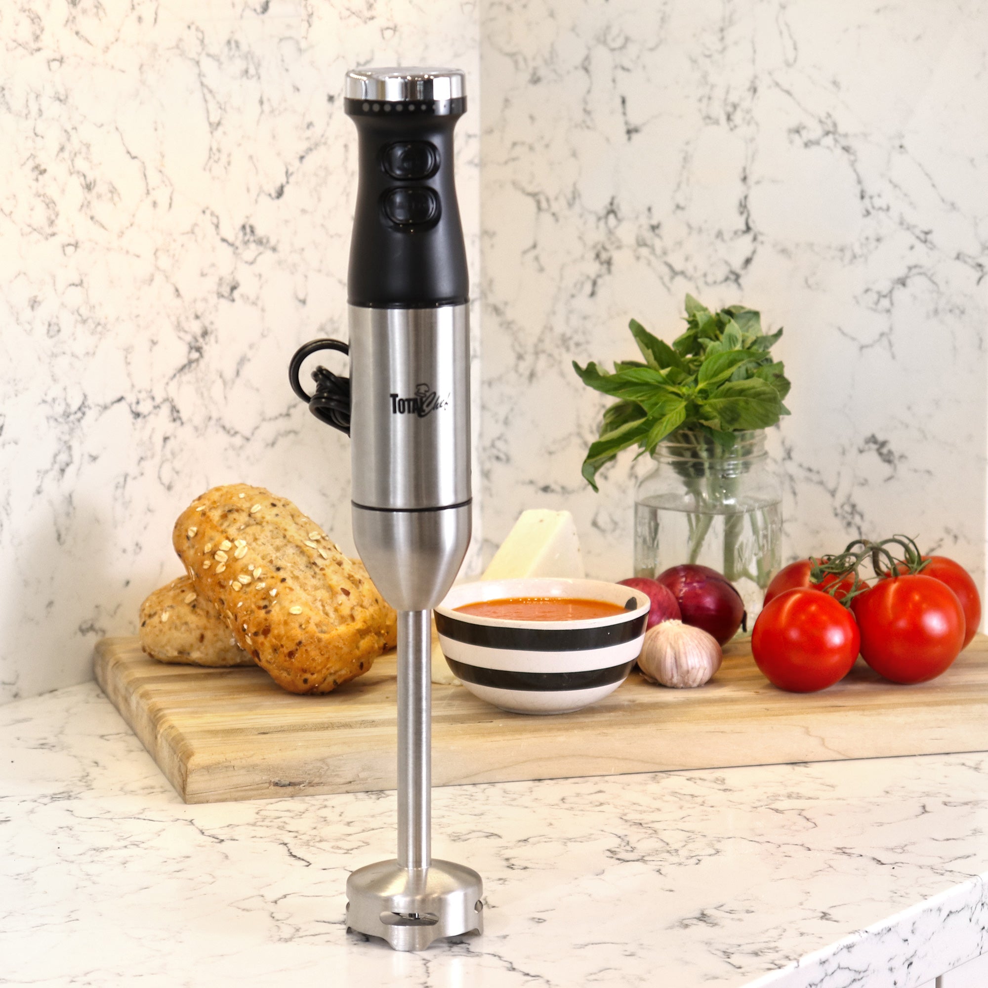 Lifestyle image of blender standing upright on a marbled white counter in front of a wooden cutting board with small loaves of bread, a bowl of soup, tomatoes, garlic, and basil behind it