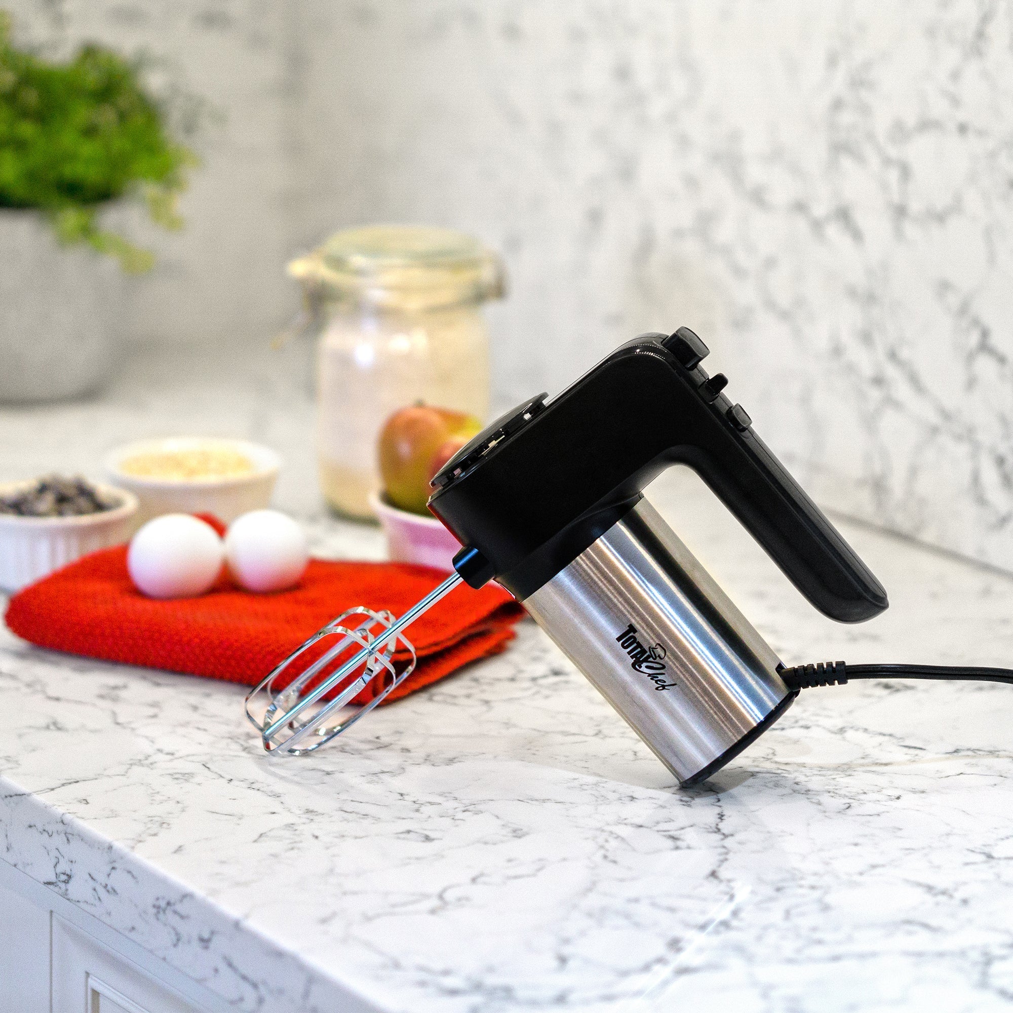 Lifestyle image of mixer on marbled white countertop with red dish towel, eggs, and other baking ingredients in the background