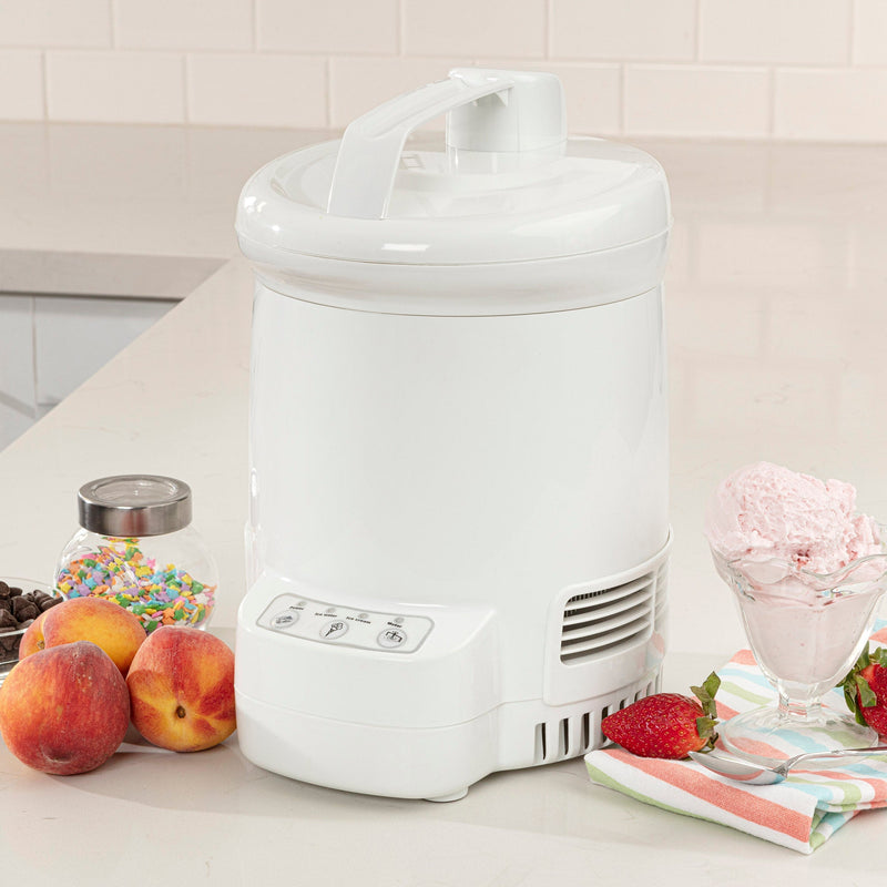 Lifestyle image of ice cream maker on a white counter with a cup of light pink ice cream, strawberries, a spoon, and a striped napkin to the right and peaches, a jar of sprinkles, and a bowl of chocolate chips to the left.