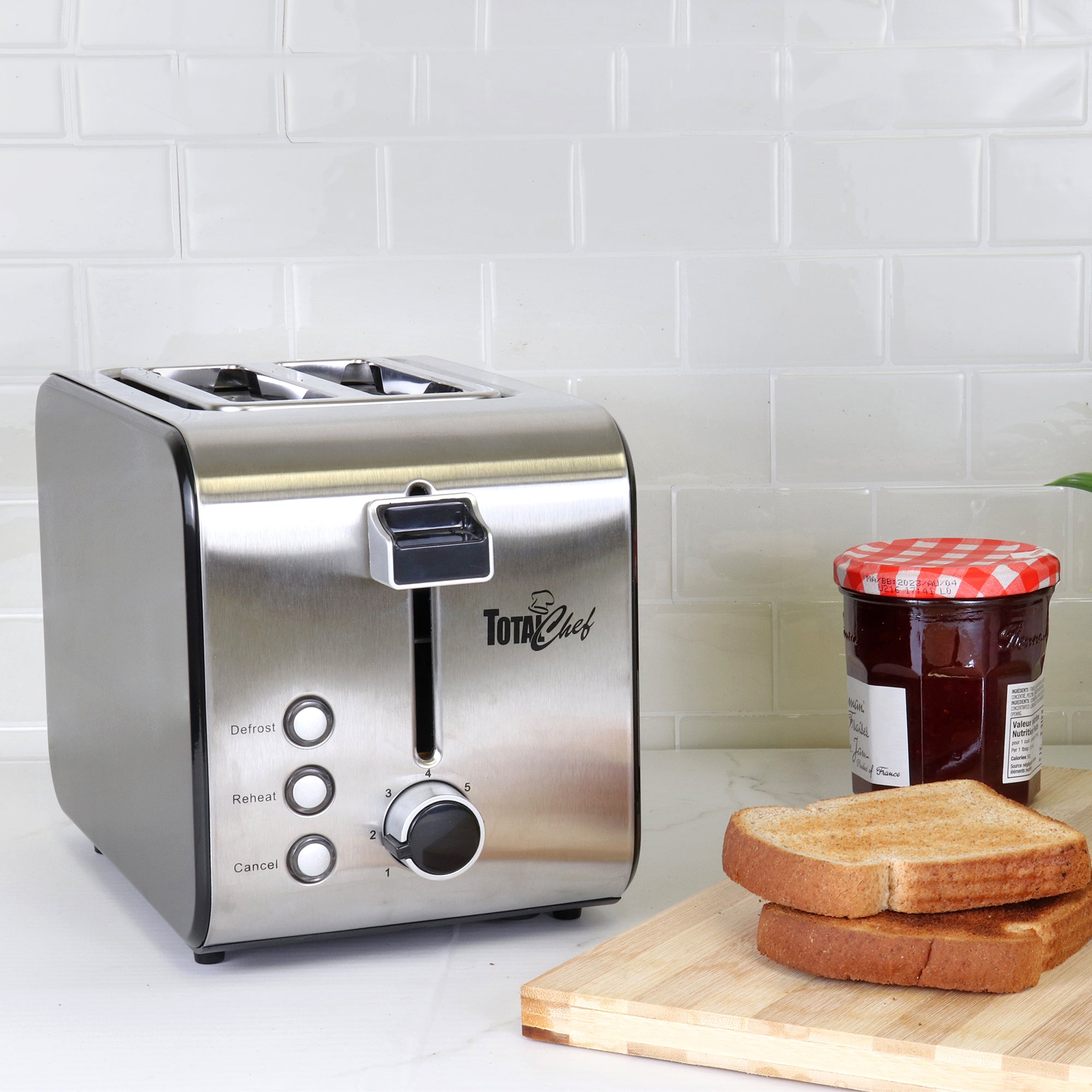 Lifestyle image of toaster beside cutting board with two slices of toast and a jar of jam on a white countertop with white backsplash