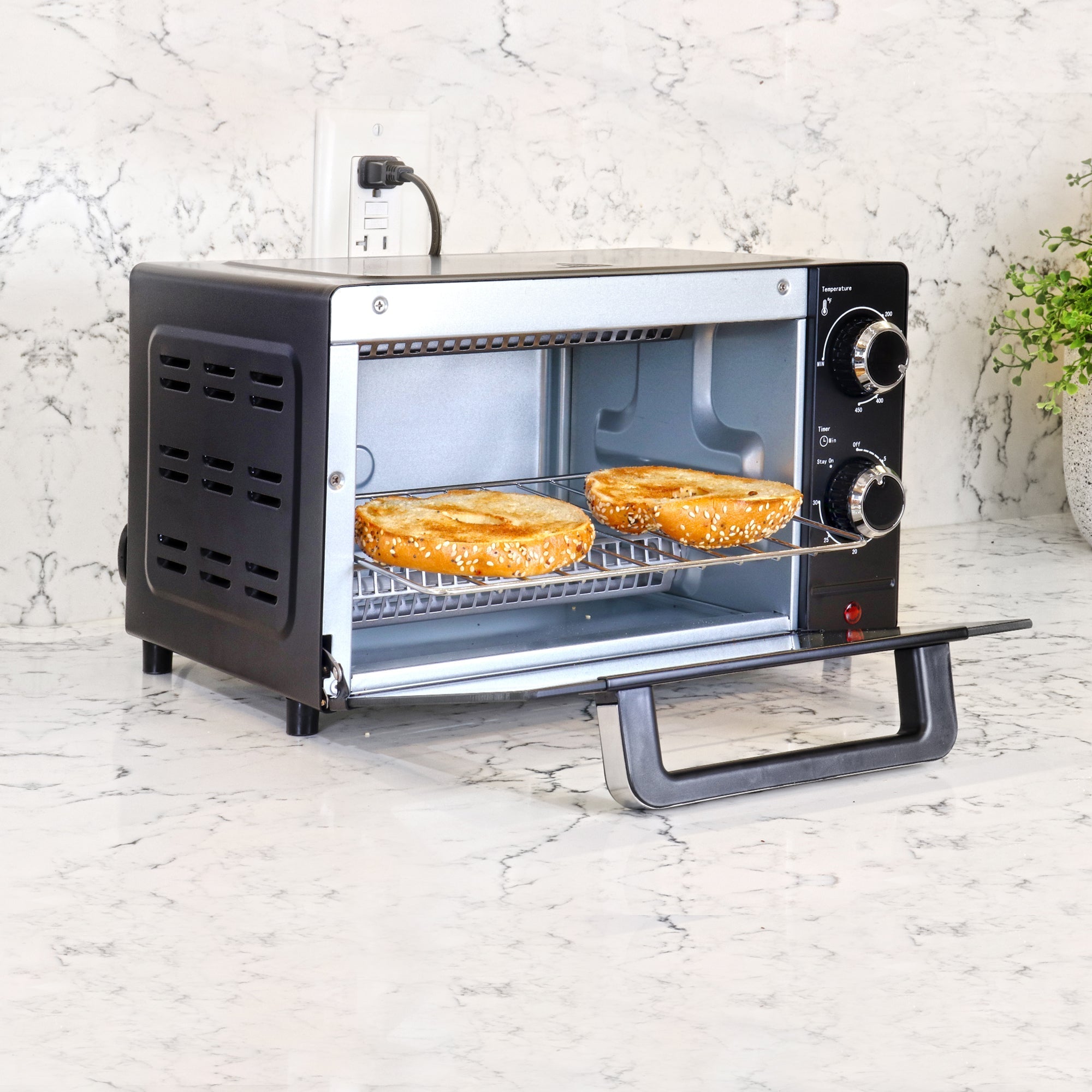 Lifestyle image of toaster oven with door open and two toasted bagel halves inside on a white marble counter with a white marble backsplash 