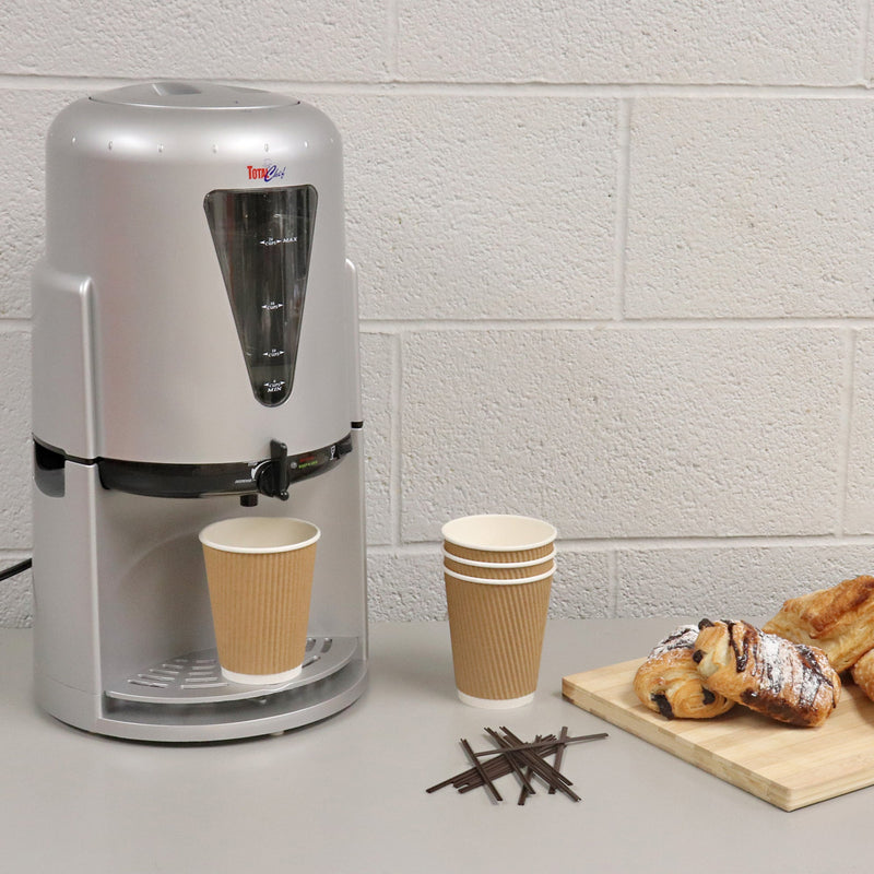 Lifestyle image of Total Chef 24 cup drink warmer on a light gray table with a white painted brick wall behind. There is a brown disposable coffee cup under the dispenser spout and a stack of cups, a pile of coffee stirrers, and a wooden cutting board with chocolate croissants to the right