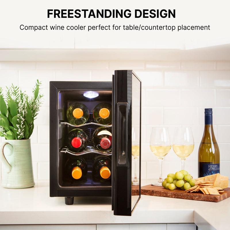 Koolatron 6 bottle countertop wine chiller, open and filled with bottles of wine, on white kitchen counter with a light green vase of wildflowers to the left and a bottle and glasses of wine, green grapes, and crackers on a cutting board to the right. Text above reads "Freestanding design: Compact wine cooler perfect for table/countertop placement"