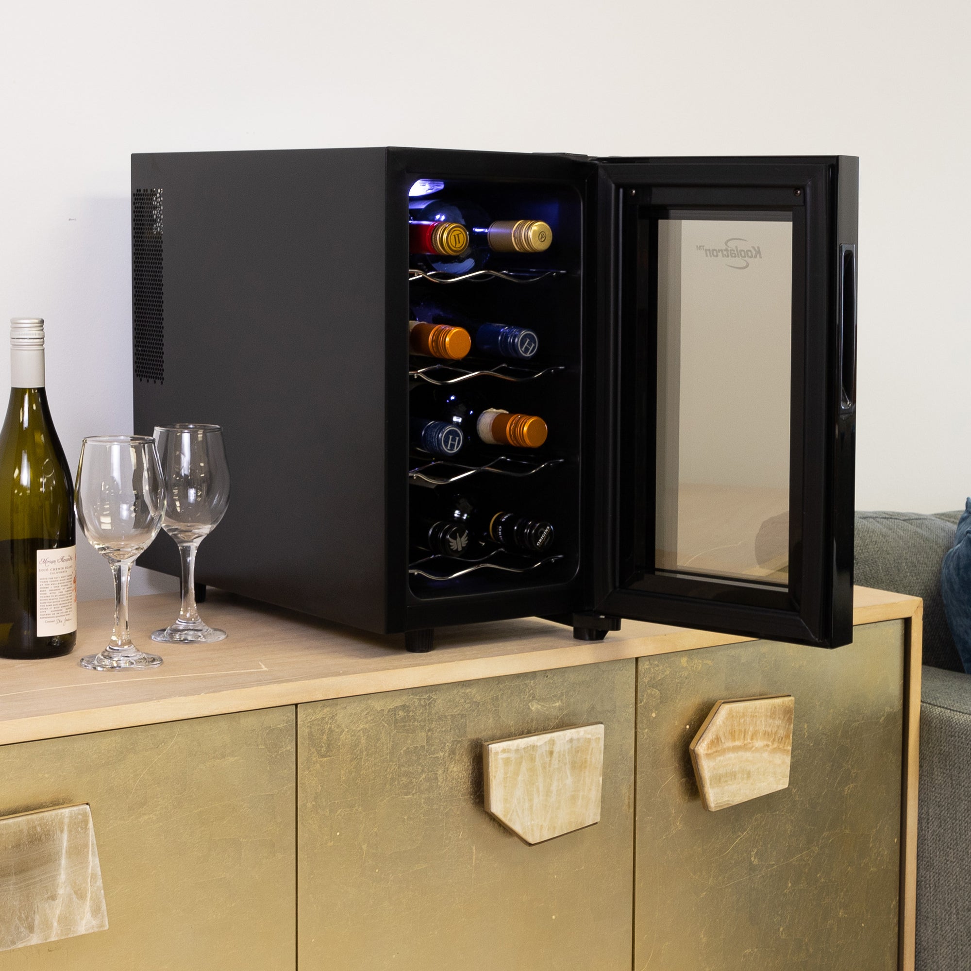 Koolatron 8 bottle wine cooler, open and filled with bottles of wine, on a gold-colored sideboard beside a gray sofa. There is a bottle of wine and two glasses to the left of the cooler