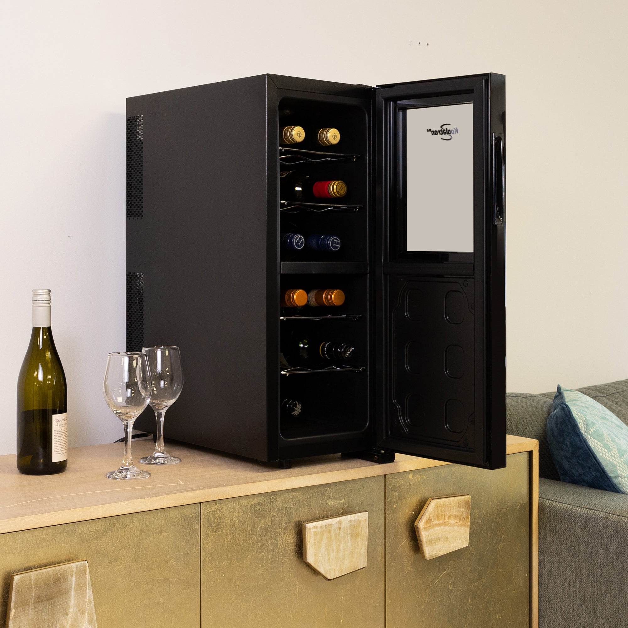 Koolatron 12 bottle wine cooler, open and filled with bottles of wine, on a gold-coloured sideboard with a bottle of wine and one glass beside it