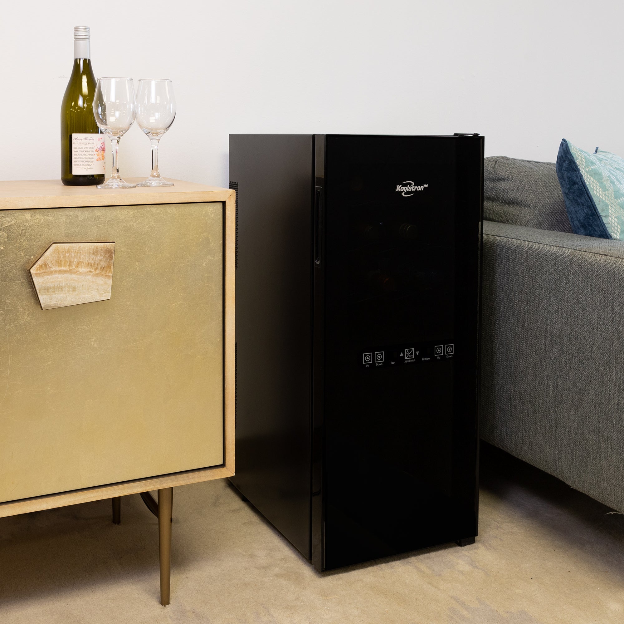 Koolatron 24 bottle dual zone wine cooler, closed, between a gray sofa and a gold-coloured sideboard with a bottle of wine and two glasses on it