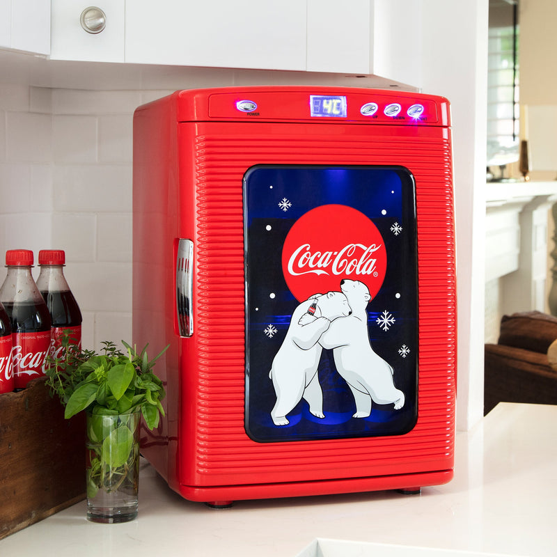 Lifestyle image of Coca-Cola 28 can mini fridge with display window, closed with blue interior light on, on a white countertop with a white tile backsplash behind it and bottles of Coke and a vase of flowers to the left