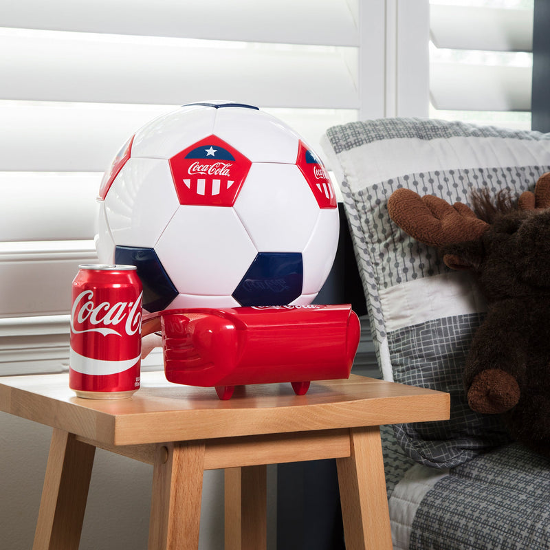 Lifestyle image of Coca-Cola soccer ball shaped mini fridge and can of Coke on a light-colored wooden bedside table. There is a bed with gray and white striped bedding and a stuffed moose to the right and a window with white wooden blinds behind