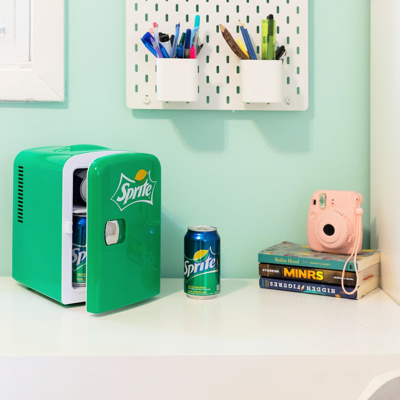 Lifestyle image of Coca-Cola Sprite 6 can mini fridge, partly open with cans inside, on a white desktop with an aqua wall behind. There is a can of Sprite, a stack of books, and a pink camera to the right of the fridge and two cups of pens and pencils attached to the wall above