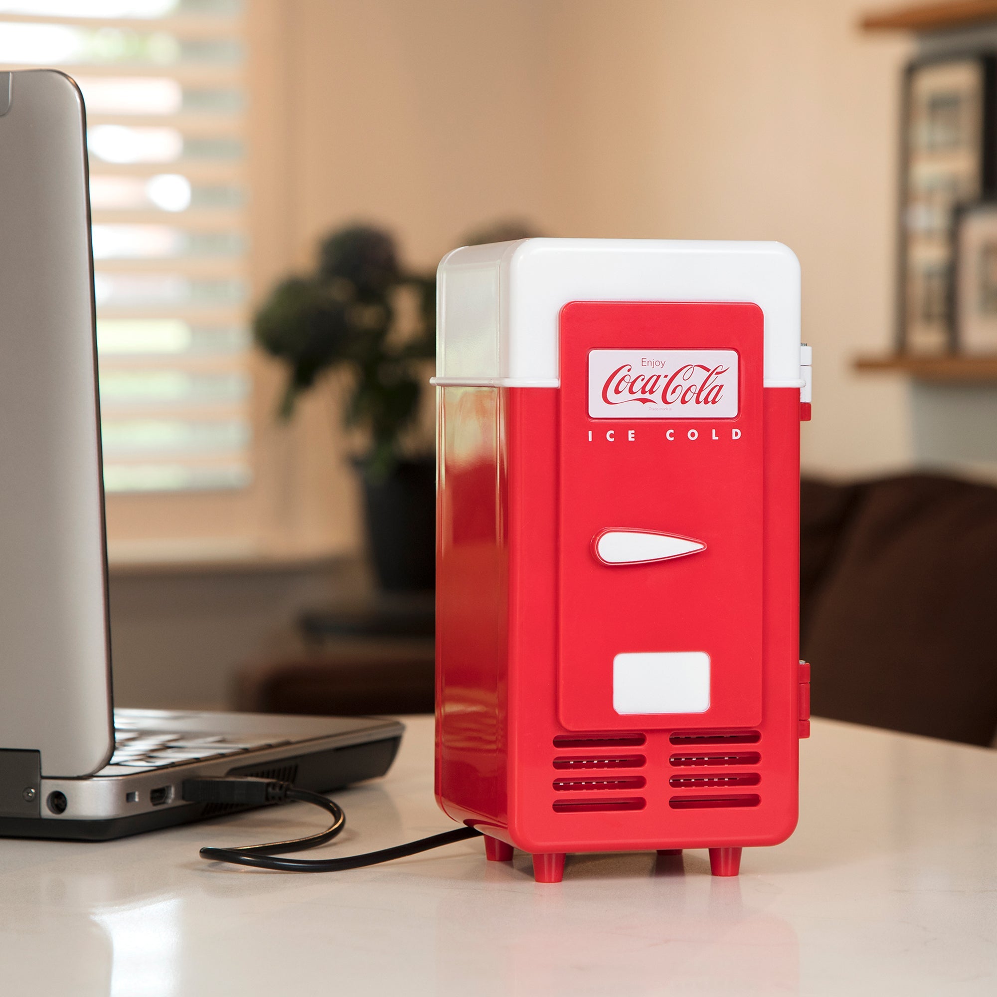 Lifestyle image of Coca-Cola single can USB cooler, closed, plugged into a silver-gray laptop computer on a light beige desktop