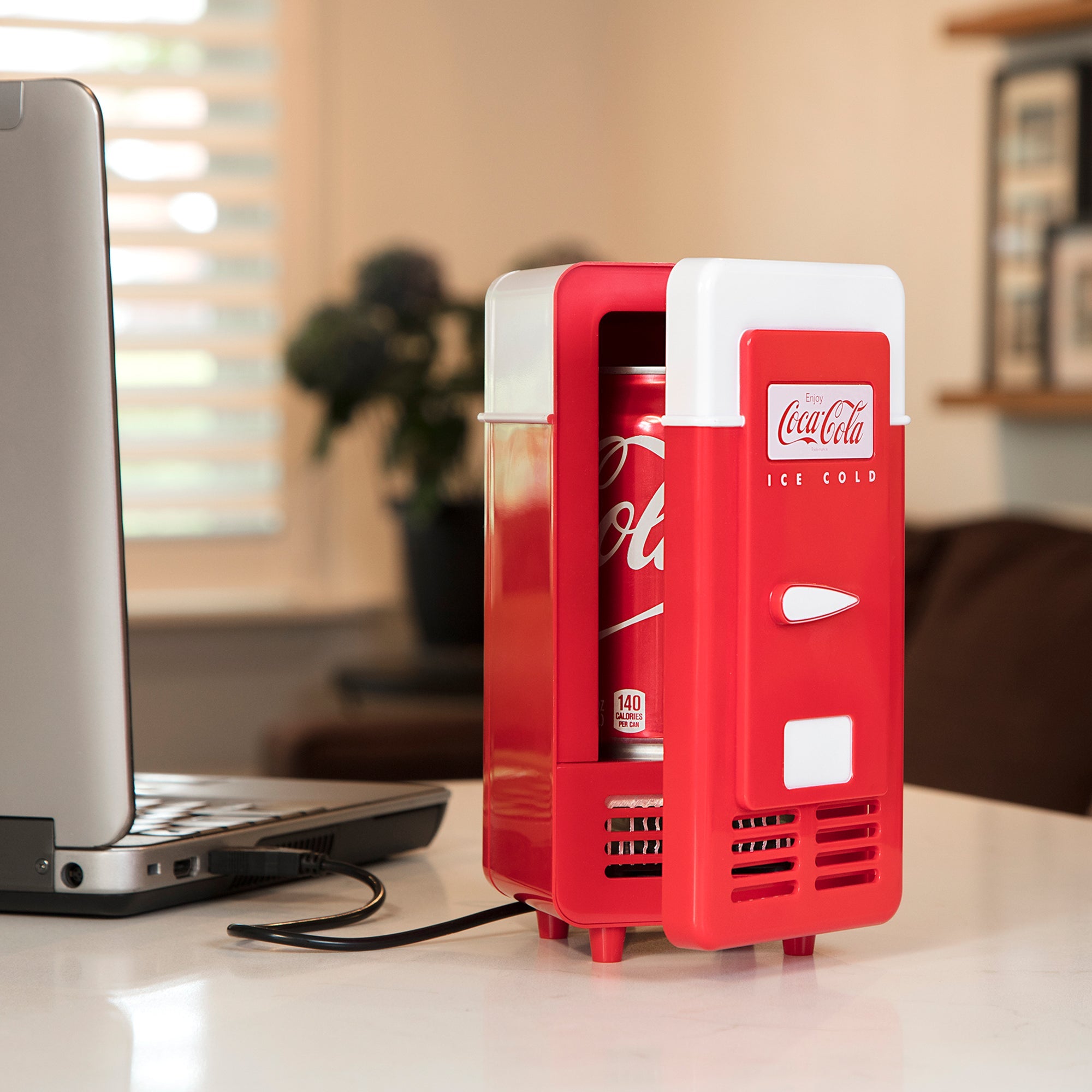 Lifestyle image of Coca-Cola single can USB cooler plugged into a silver-gray laptop computer on a light beige desktop. The mini fridge is partly open with a can of Coke visible inside