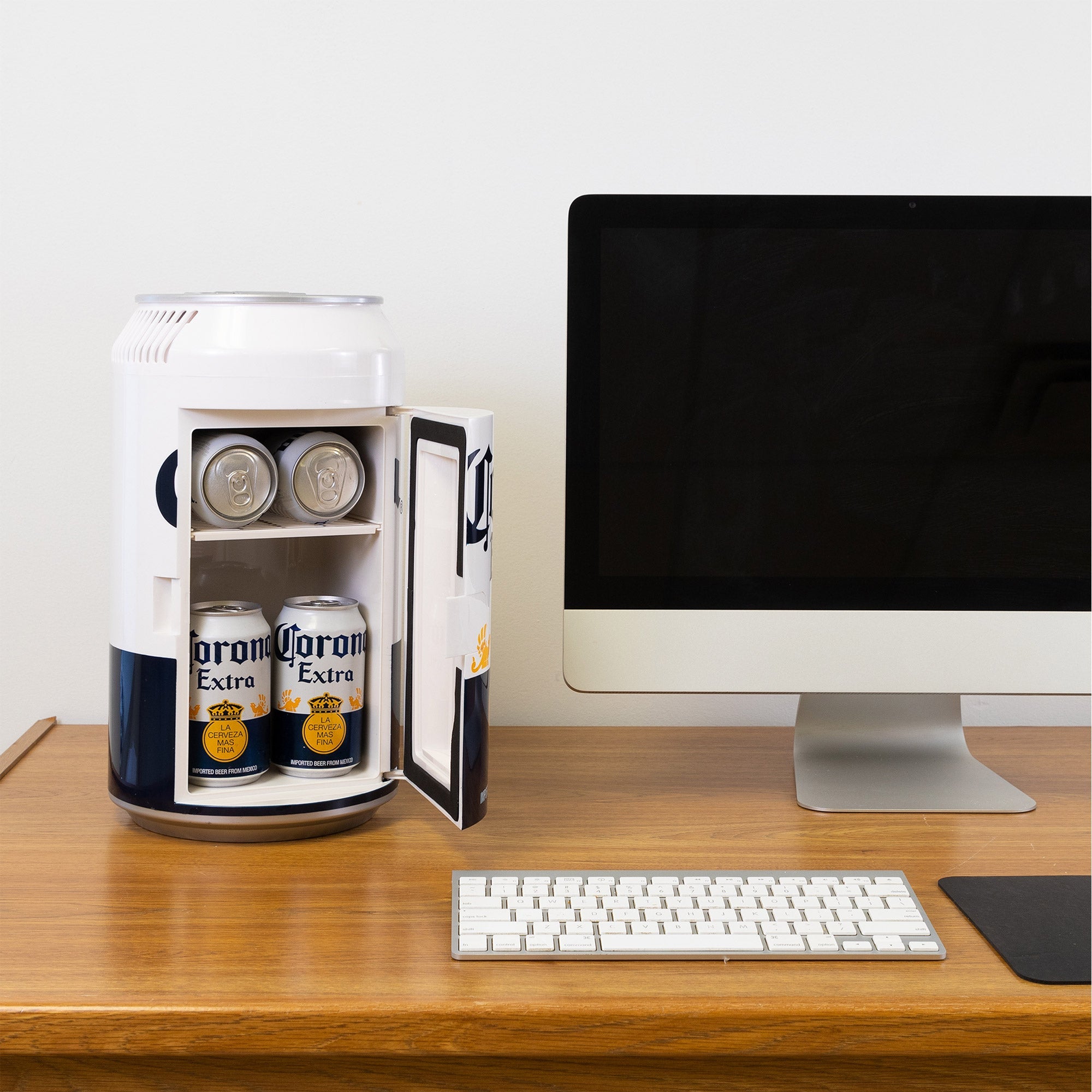 Lifestyle image of Corona can-shaped mini fridge, open with 6 cans of Corona beer inside, on a wooden desktop with a computer monitor and keyboard beside it