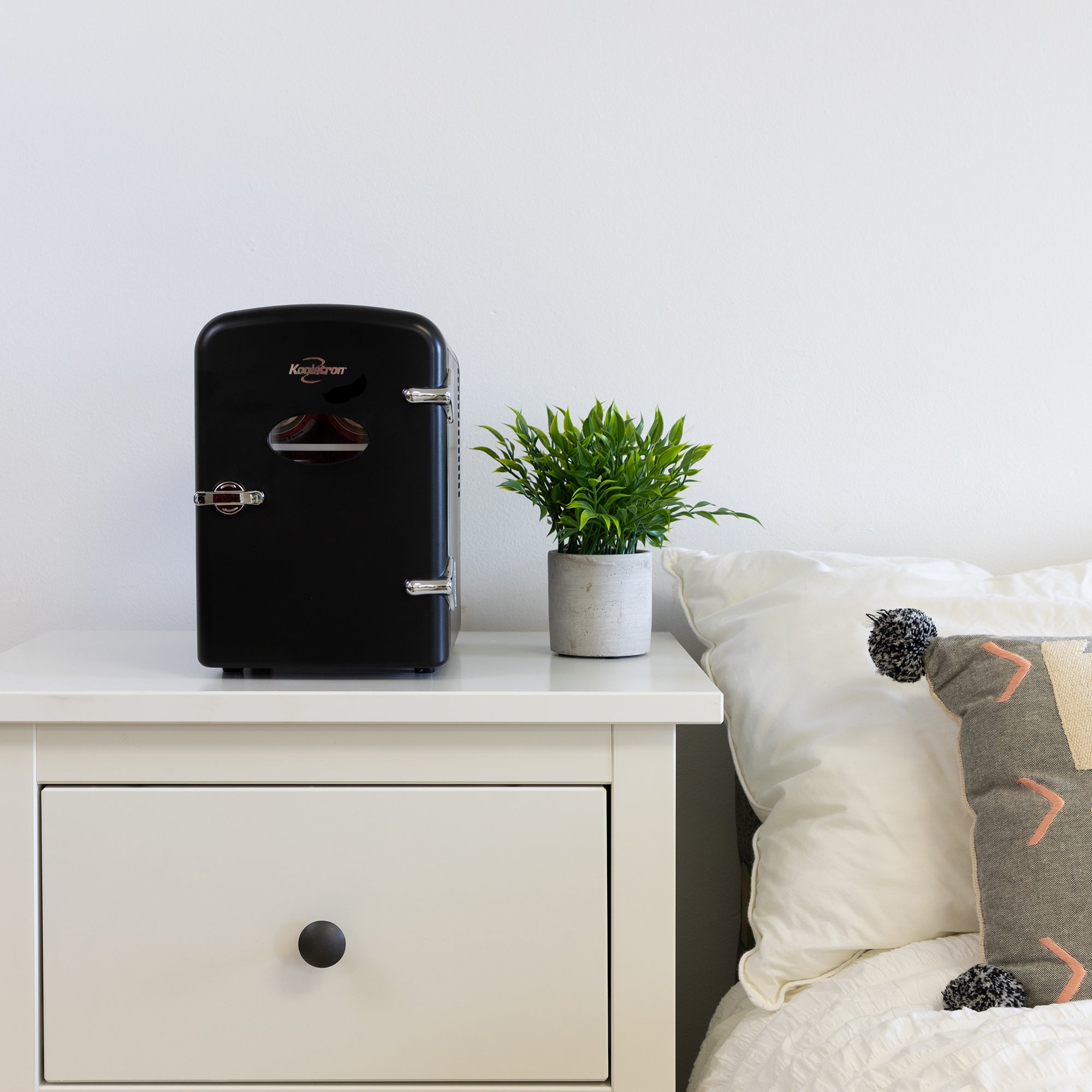 Lifestyle image of Koolatron retro 6 can mini fridge, closed, with a plant in a white pot on its right, on a white bedside table beside a bed with white linens 