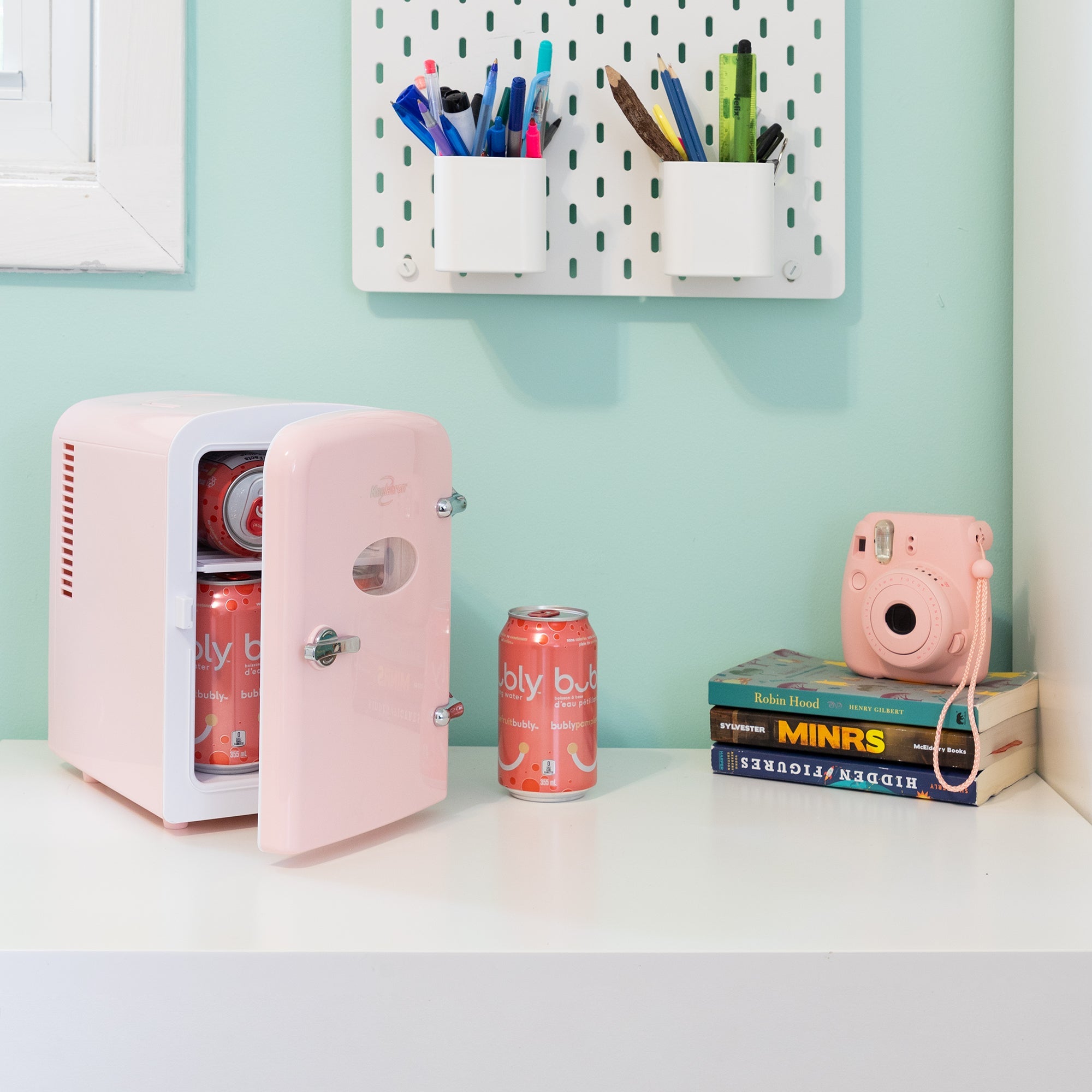 Lifestyle image of Koolatron retro 6 can mini fridge, partly open with cans inside, on a white desktop with an aqua wall behind. There is a can of soda, a stack of books, and a pink camera to the right of the fridge and two cups of pens and pencils attached to the wall above