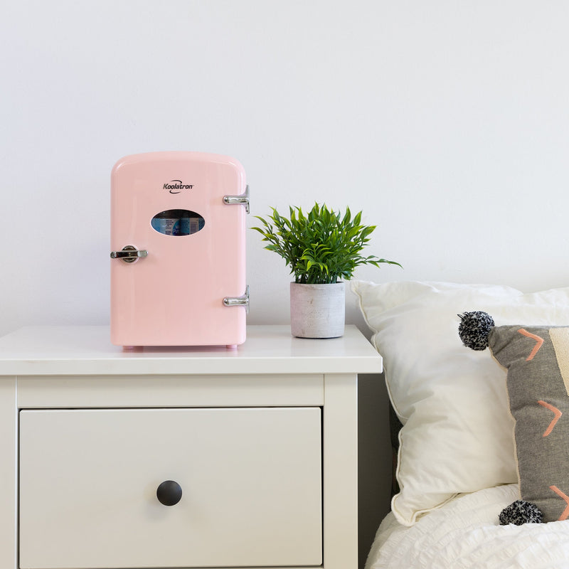 Lifestyle image of Koolatron retro 6 can mini fridge, closed, with a plant in a white pot on its right, on a white bedside table beside a bed with white linens
